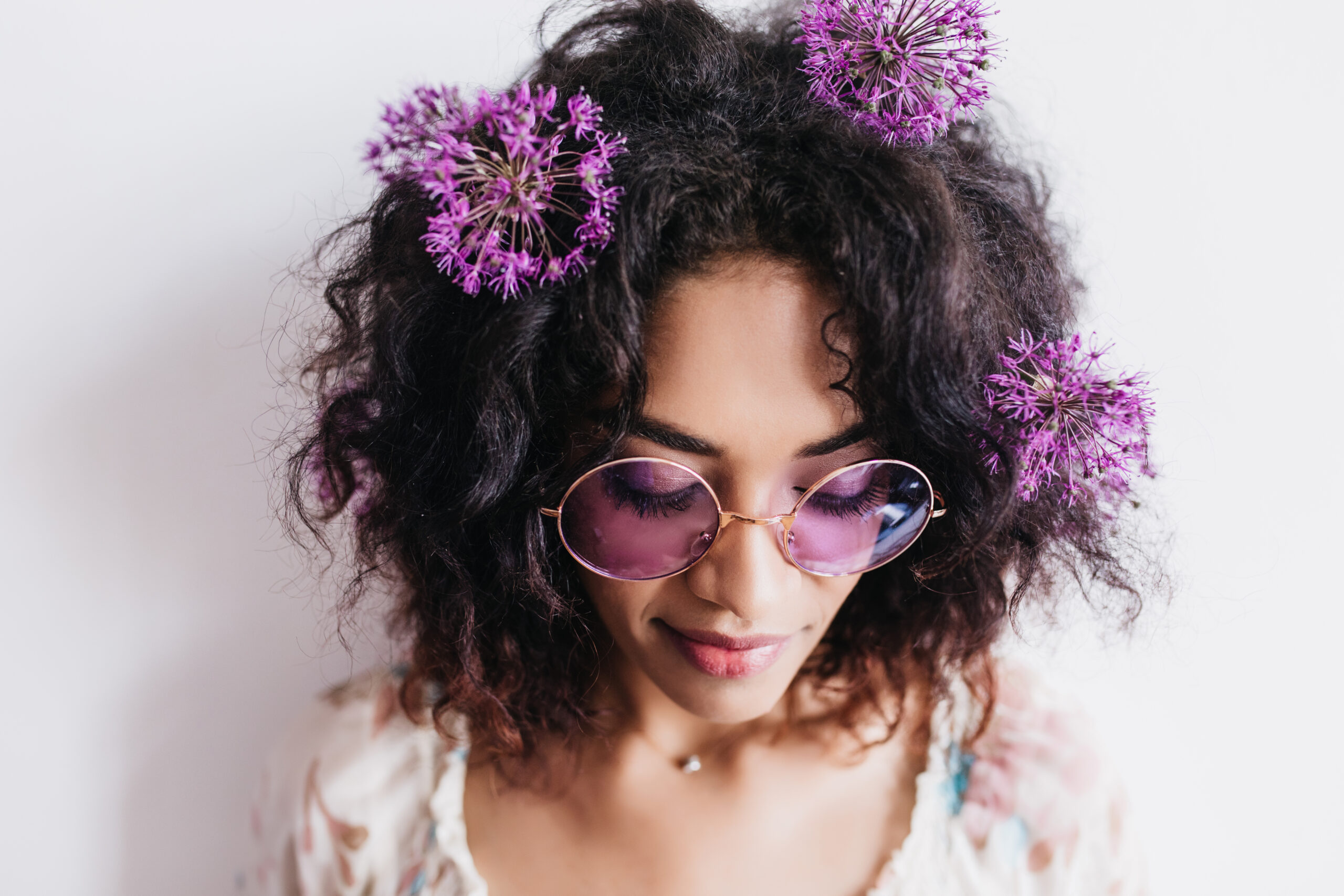 Close-up portrait of pretty african woman in sunglasses posing with eyes closed. Studio shot of wonderful female model with dark hair isolated on white background with purple flowers.